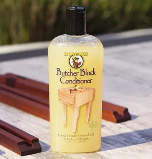 A 12 oz. bottle of Howard Butcher Block Conditioner is sitting atop a grey table top, with sunlight shining down on it from the left. Wooden jigs are in the background.