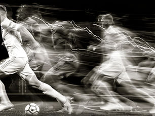 A black and white photo of a group of FIFA soccer players running while kicking a white soccer ball on a soccer field and while wearing a white soccer uniform.