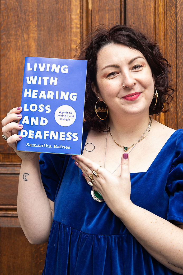 Samantha Baines holds her book and smiles at the camera. She is a white woman with short black curly hair. She has a nose ring, brown eyes, hoop earrings and a nose piercing. She is wearing a dark blue dress which matches her book cover.