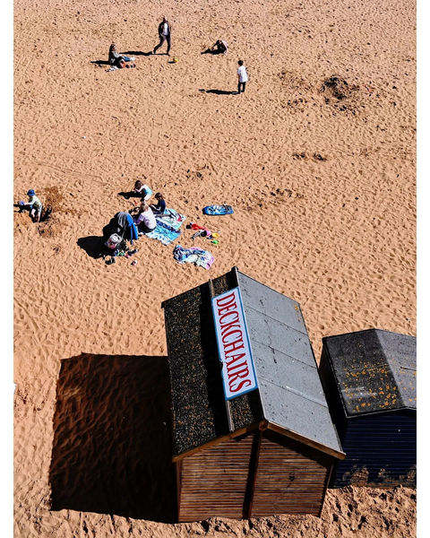 Wooden beach huts, DECKCHAIRS sign, people relaxing on sunny sand.