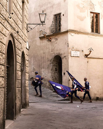 People in historical attire carrying blue banners on a narrow European street.