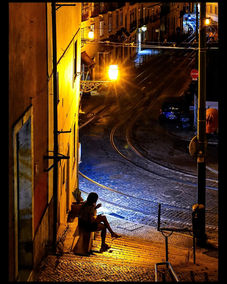 Person sitting on dark cobblestone street at night, lit by old streetlamp.