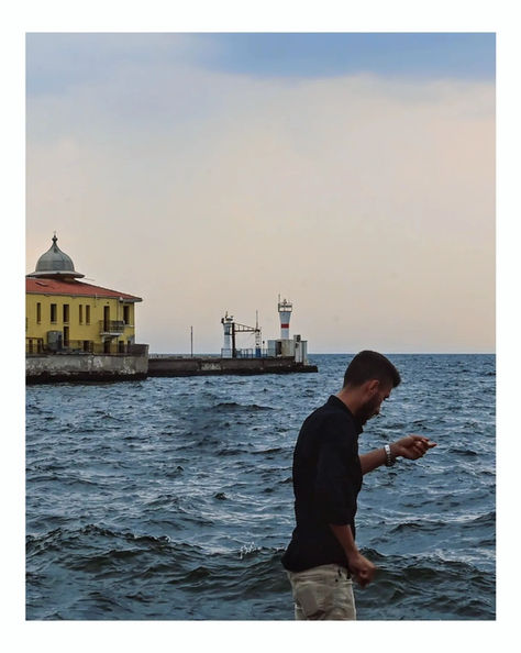 Man looking at phone by wavy sea, historic building in background.