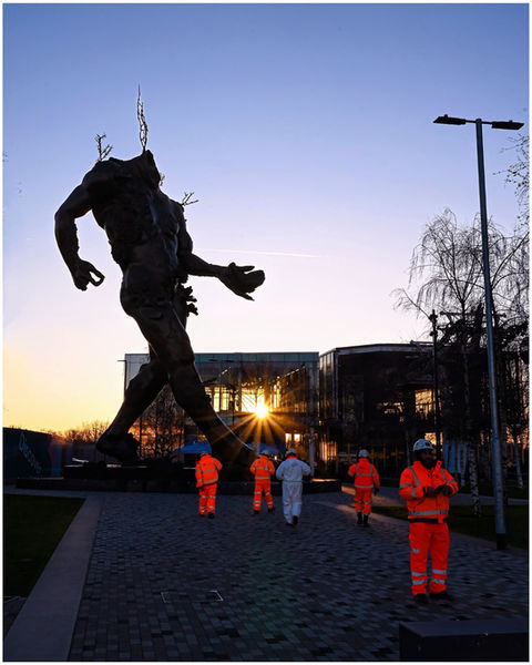 Massive human figure sculpture, workers in orange high-visibility suits at sunset.
