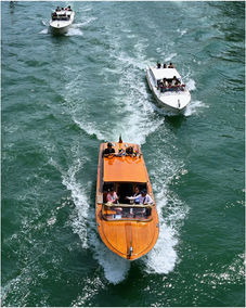Orange wooden boat leads two white speedboats on a wavy green channel.