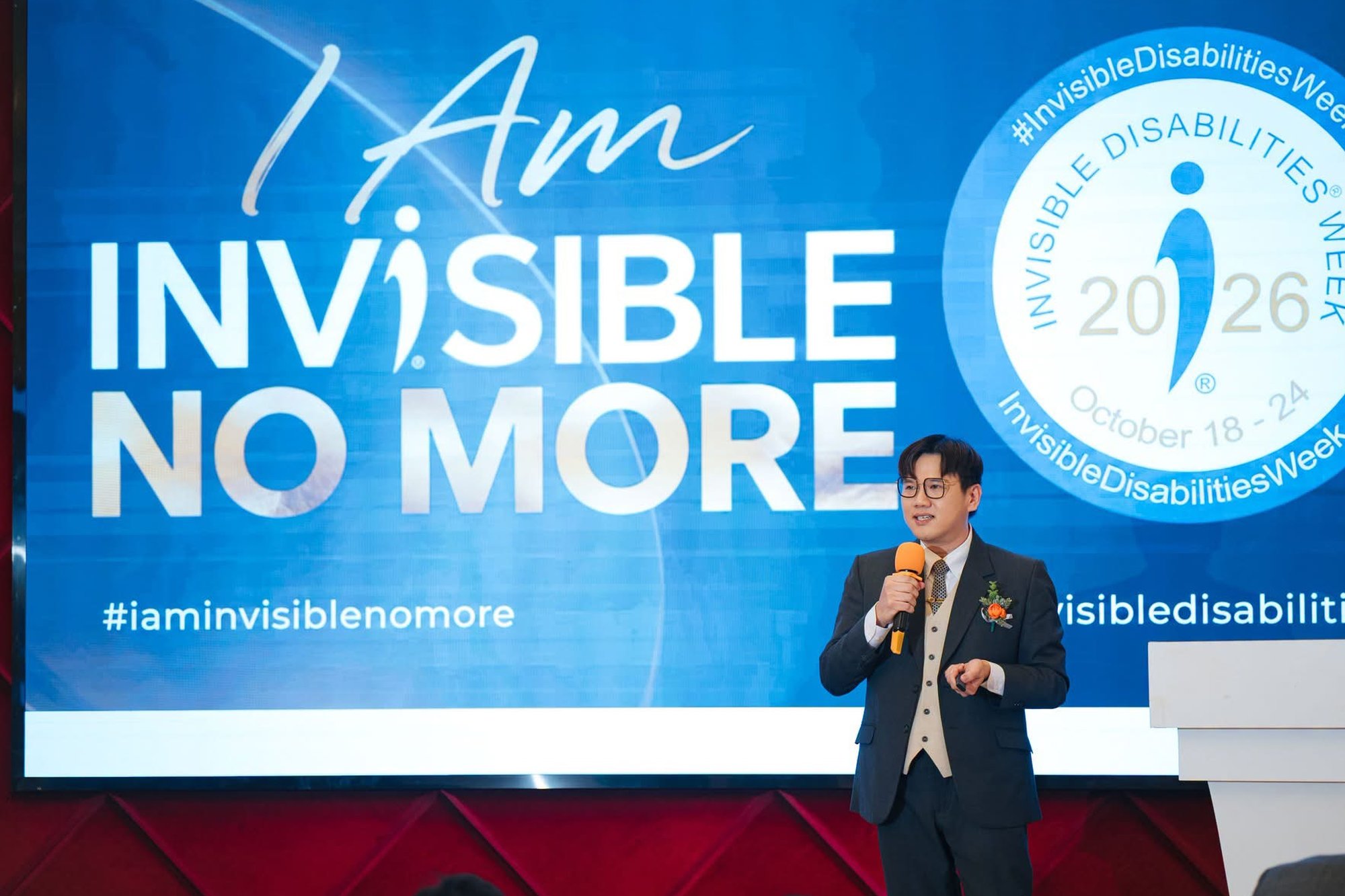 Speaker on stage with opening slide reading I Am Invisible No More, Invisible Disabilities Week 2026.