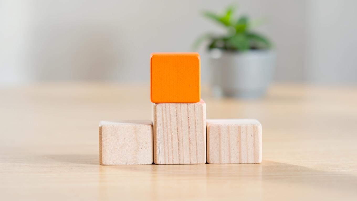 Five plain wooden blocks arranged as ascending steps on a desk, the tallest painted bright orange, a plant blurred behind