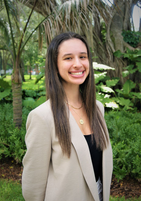 Woman with long straight brown hair with highlights, and wearing two gold necklaces, a beige blazer, and a black shirt