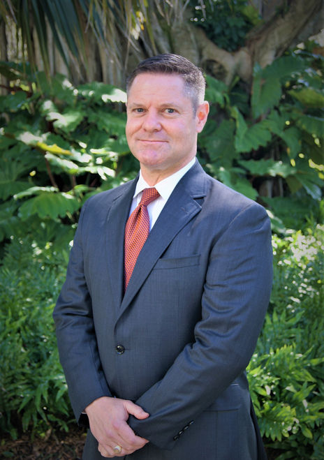 White man with short brown and white hair, and wearing a dark grey blazer, white button-down shirt, and an orange tie