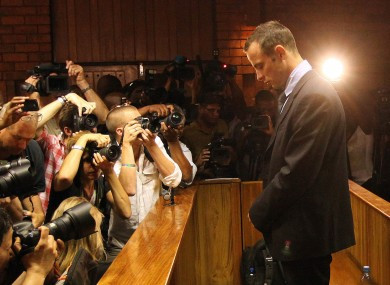 Oscar Pistorius stands with his head bowed, dressed in a dark black suit and blue shirt. On the left, a crowd of photographers are taking photographs