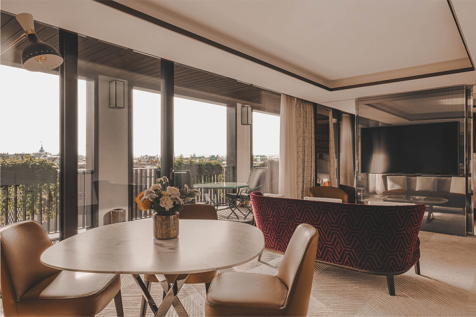 Elegant living room with a round table, beige chairs, and a red-patterned sofa. Large windows reveal a balcony view. Warm, cozy ambiance.