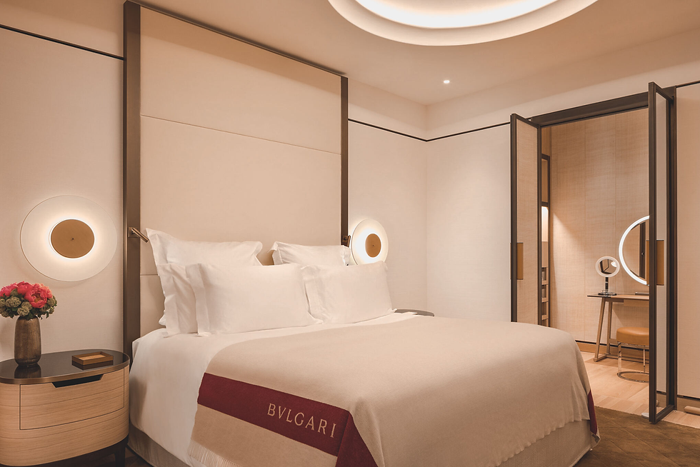 Elegant hotel room with a plush bed, white linens, and a BVLGARI blanket. Warm lighting, a flower vase, and a vanity with a round mirror. Cozy ambiance.