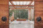 Open wooden doors reveal a serene patio with wicker chairs, a wooden bench, and plants. Sunlight filters through a geometric lattice.