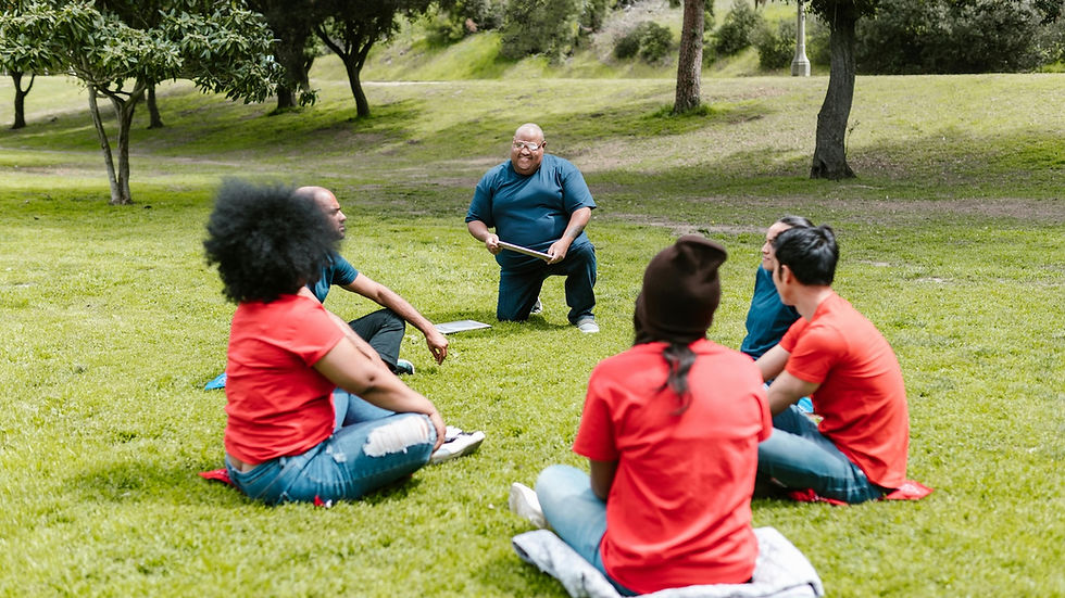 A group of five people sit on the grass in a park, listening to a person kneeling with a notebook. Trees and greenery in the background.