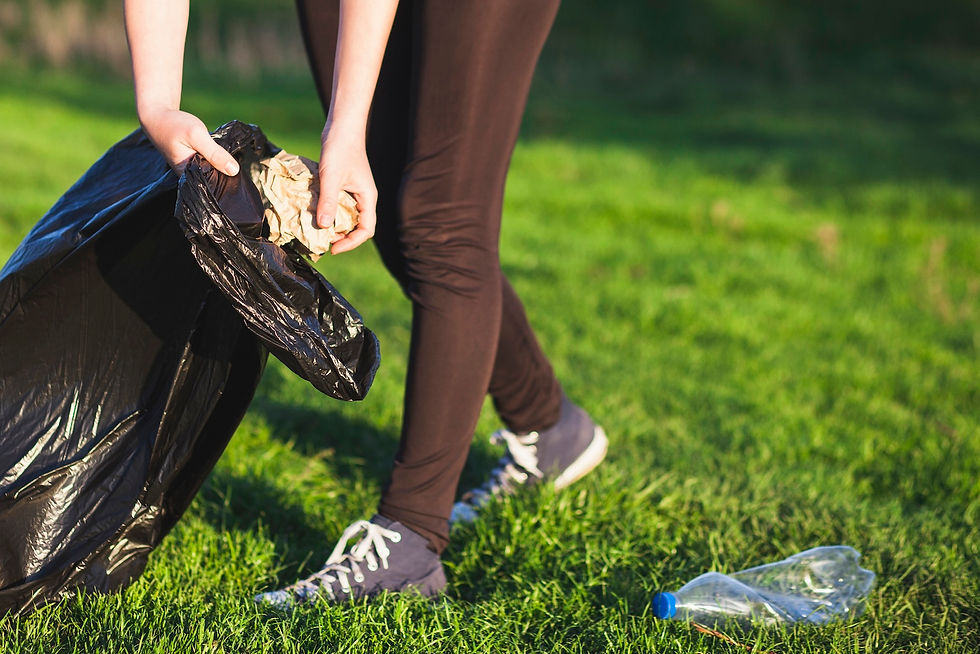 Person in black pants picks up trash from green grass into a black bag. A crumpled plastic bottle lies nearby. Outdoor cleanup scene.