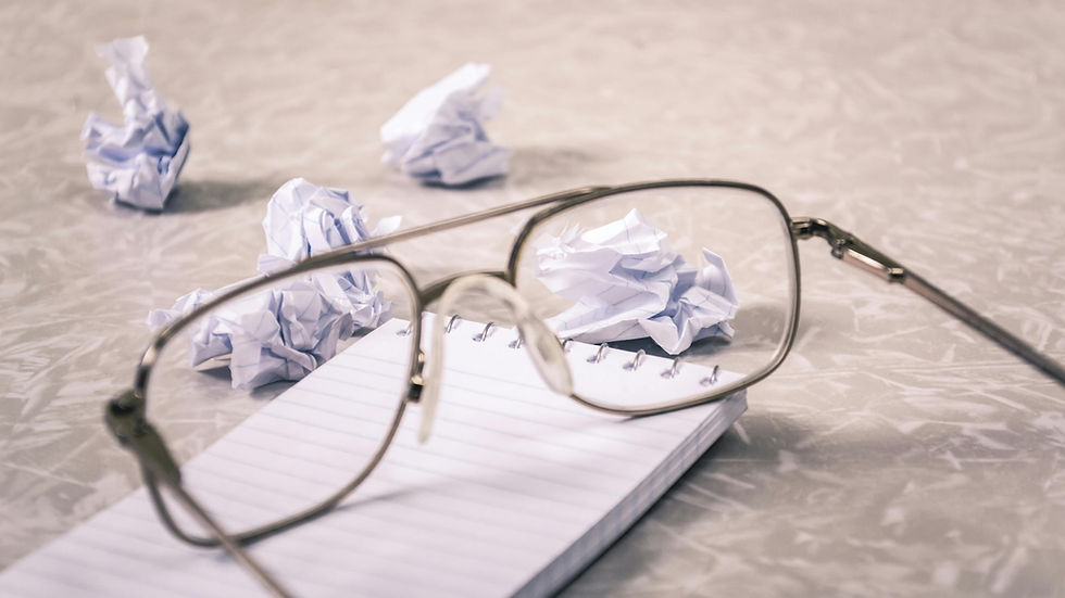 A pair of eyeglasses resting on a lined notepad with several crumpled pieces of paper scattered on a desk, symbolizing the frustration of revising and reworking proposals or budgets.