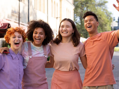 Four friends in colorful outfits laugh together on a sunny street, exuding joy and camaraderie, with trees and buildings in the background.