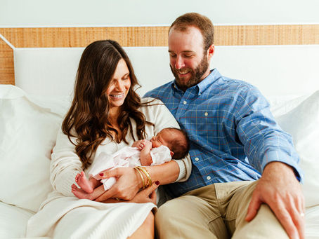 A mom and dad sitting on their bed holding baby girl smiling at her