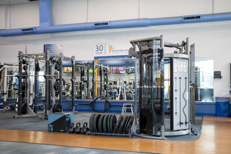 A bright and spacious gym. Multiple pieces of equipment are featured. Smith machine, bench press, squat racks and functional trainer