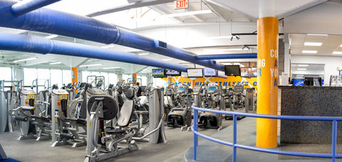 A large, brightly lit gym is pictured. Multiple rows of equipment including selectorized weight machines and cardio equipment fill the space. 