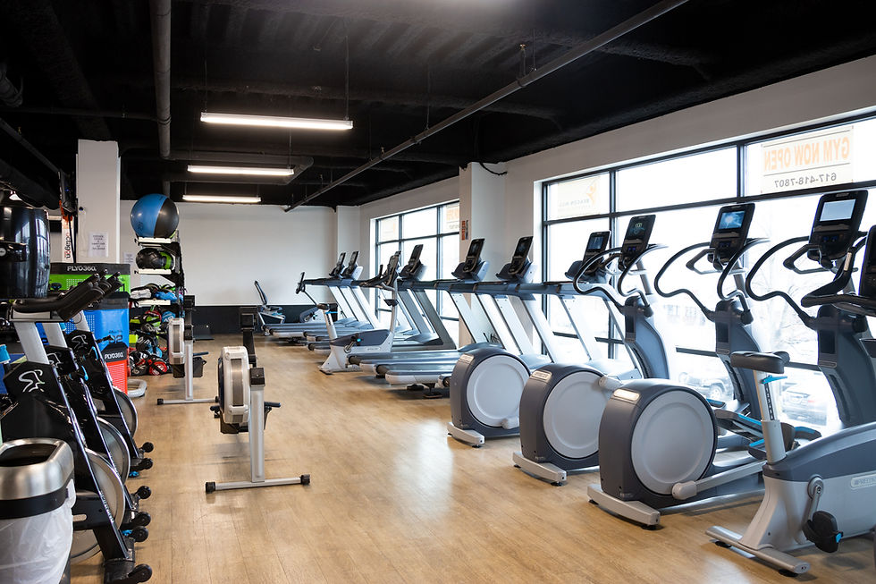 Rows of treadmills and exercise bikes in a well-lit gym with wooden floors. Bright windows display "GYM NOW OPEN" signs.