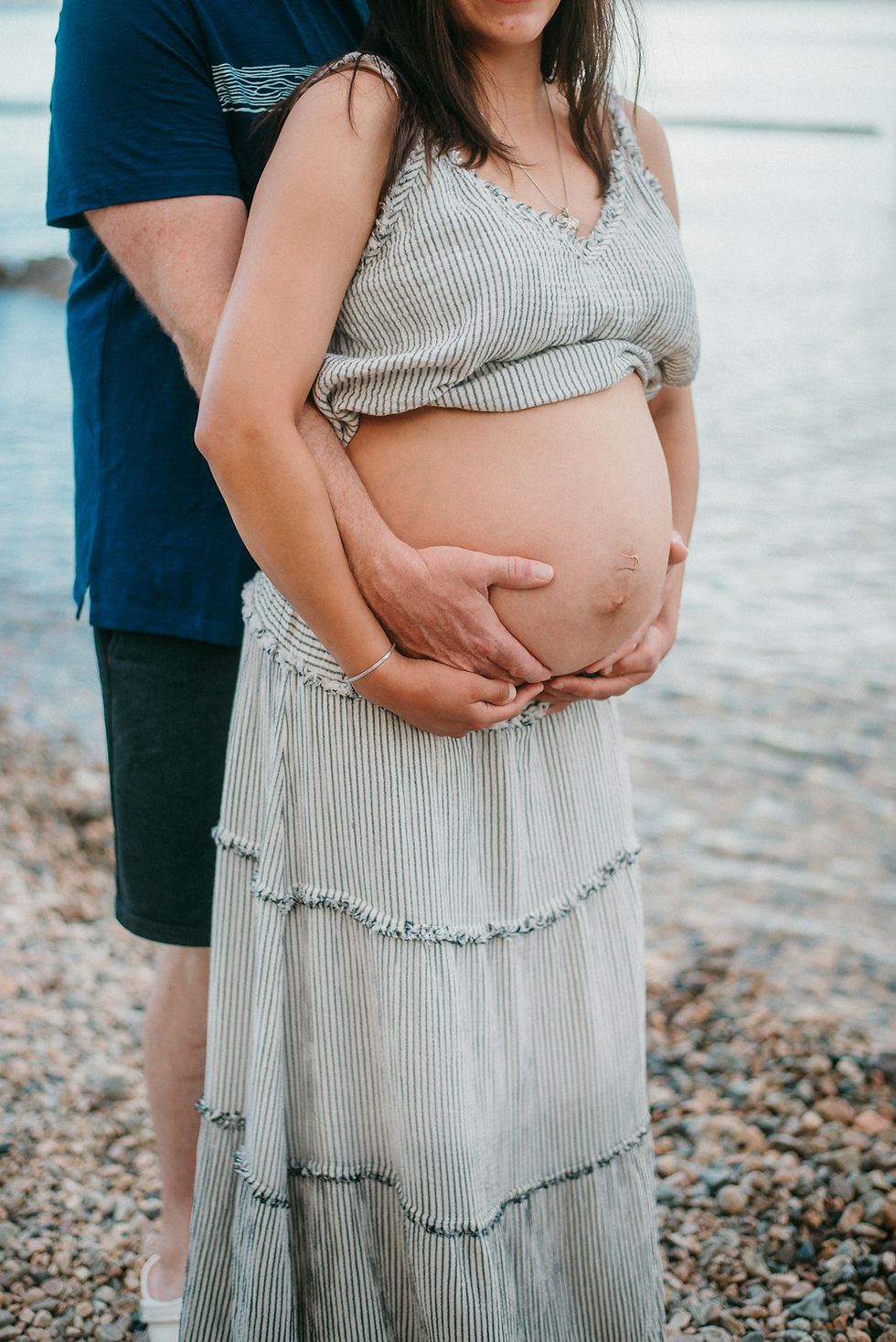 Pregnant woman in striped dress stands on rocky beach while partner hugs her baby bump from behind.
