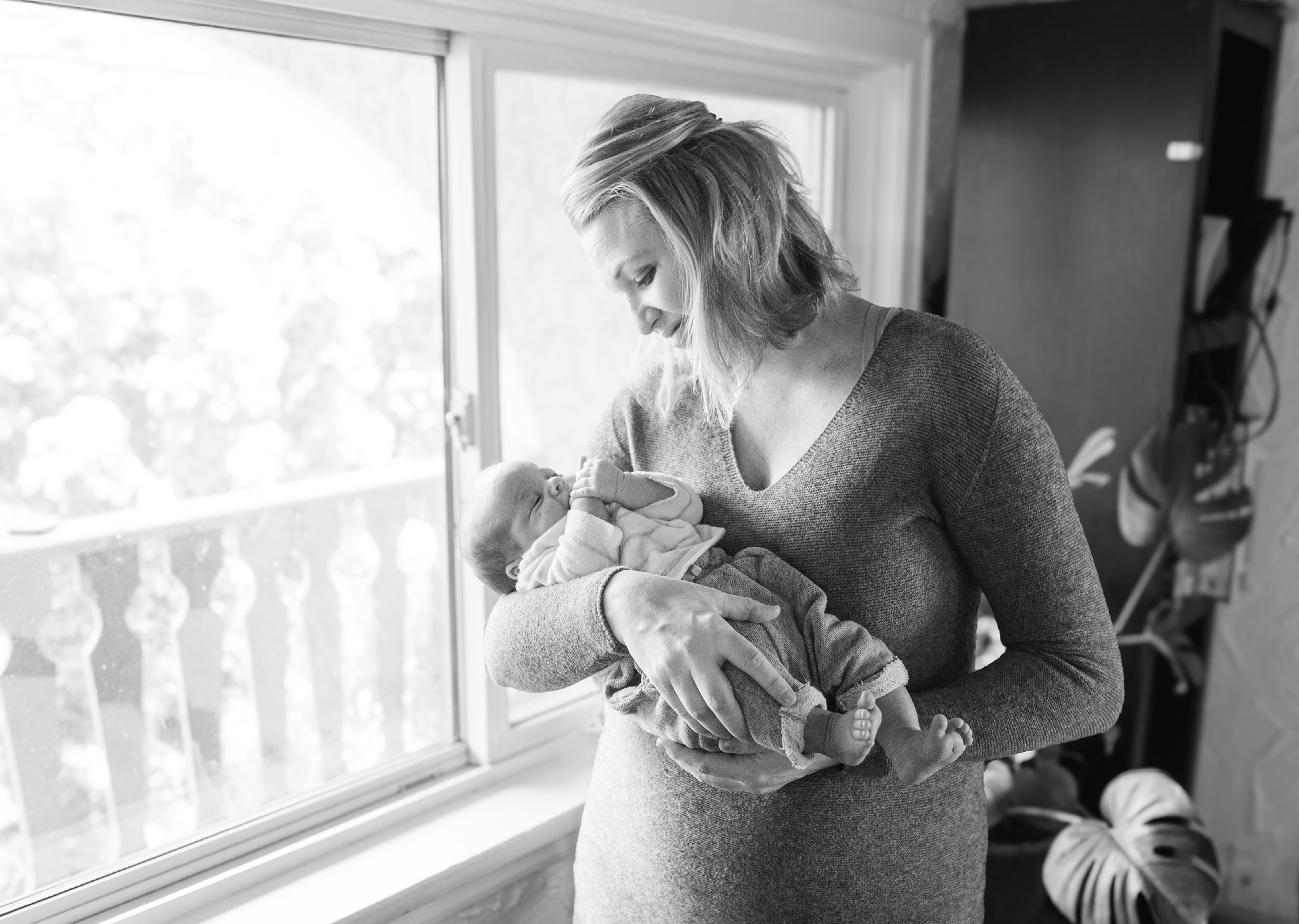 Black-and-white photo of a woman gently cradling a sleeping baby by a bright window, looking down with a tender expression.