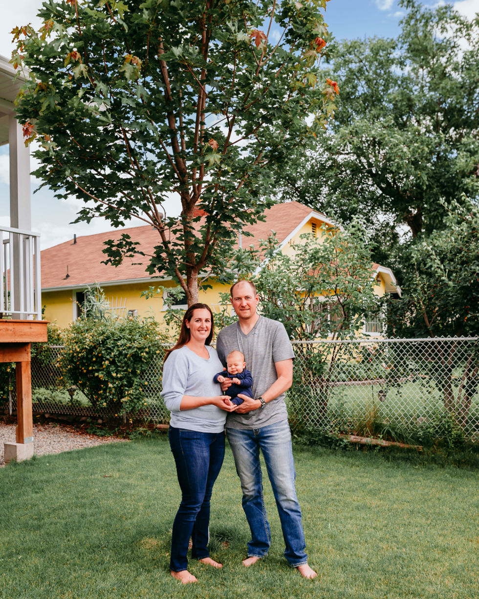 A couple sits cozily on a bed in a bright bedroom, smiling at each other with their newborn during at home photography session in Penticton