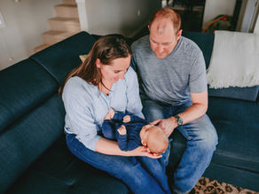 a family is sitting on the sofa for an in-home newborn photography session with their baby boy.