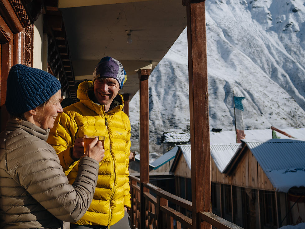 Twee bergwandelaars op een balkon in de sneeuw, lachend met drinkbekers; op de achtergrond besneeuwde bergtoppen en hutten.