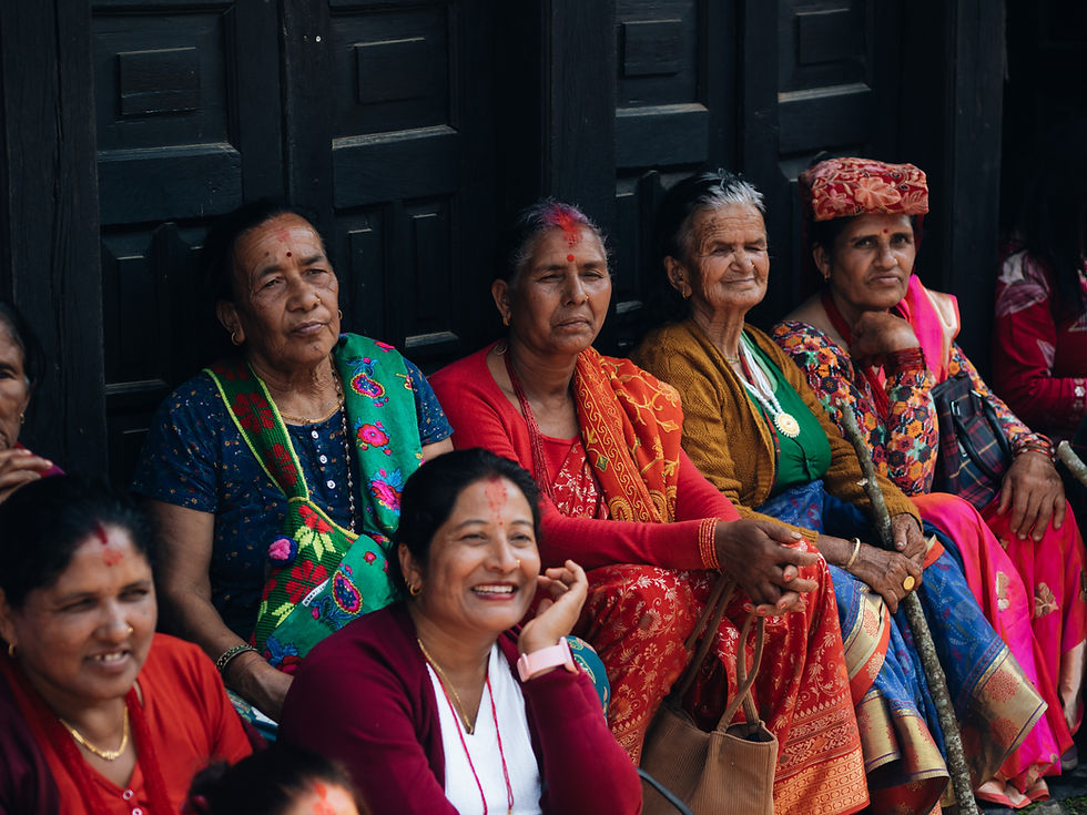 Groep Nepalese vrouwen in kleurrijke sari’s zit voor een donker houten muur; een vrouw lacht, anderen kijken ernstig.
