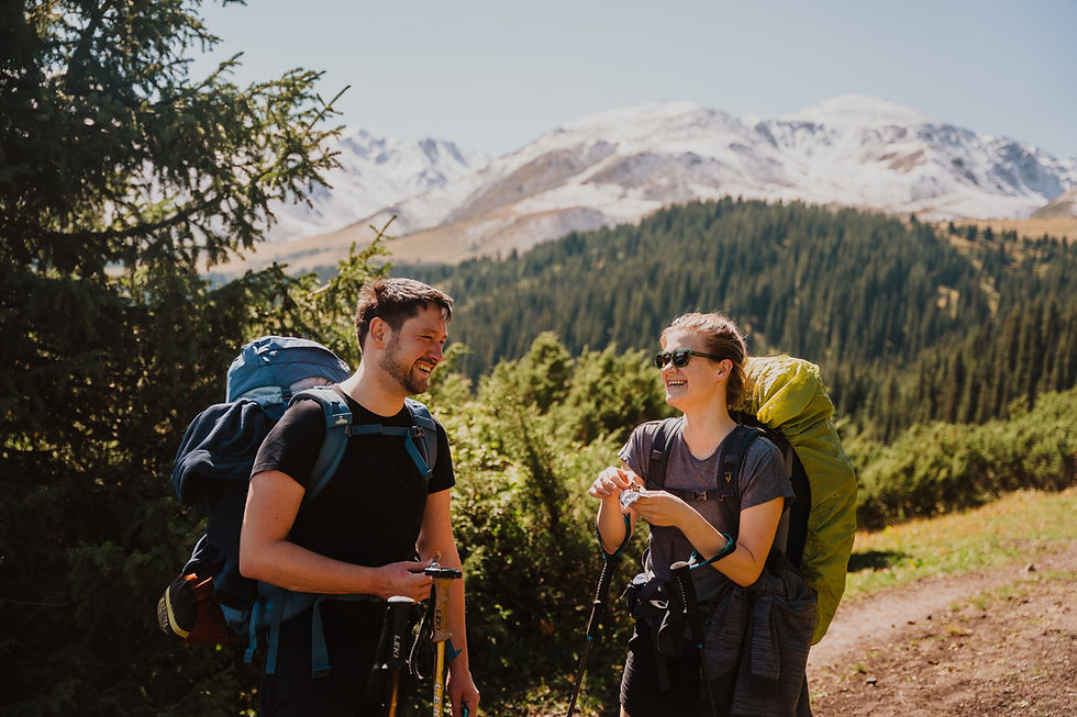 Two hikers with backpacks and trekking poles chat and smile in a mountainous forest. Sunny day, snowy peaks, and vibrant greenery.