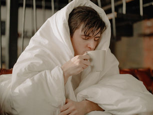 A person wrapped in a white blanket drinks from a mug, sitting on a brown leather couch in a cozy room. They appear relaxed and warm. Elderberry + Echinacea for Immunity. Herbpy Blog.