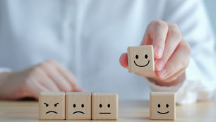 A hand picks a wooden block with a smiling face from a row of four blocks showing various emotions, set on a wooden table. Black Cohosh and St. John’s Wort for Mood Balance. Herbpy Blog.