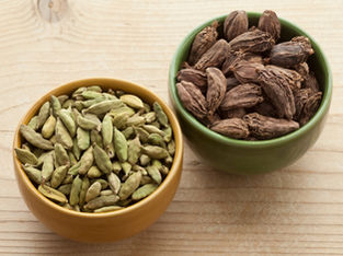 Two bowls of cardamom pods on a wooden surface; one bowl is yellow with green pods - green cardamom, the other is green with brown pods - black cardamom. Source: Canva