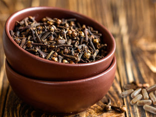 Brown bowls filled with cloves on a wooden surface. Nearby are several beige capsules, suggesting a herbal or natural theme.