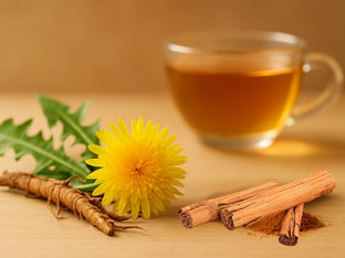 Dandelion flower, root, and cinnamon sticks on a wooden surface; a glass cup filled with golden brown tea in the background. Warm tones.