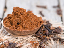 Ground cloves in a wooden bowl on rustic wooden table, with whole cloves scattered around, creating a warm, earthy feel. Source: Canva.