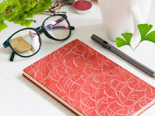 Glasses, a red patterned notebook, and a pen rest on a white table. Ginkgo Biloba tree, green Ginkgo Biloba leaves, and a pink candle create a calm setting. Source: Canva