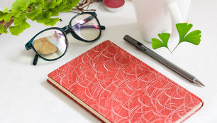 Glasses, a red patterned notebook, and a pen rest on a white table. Ginkgo Biloba tree, green Ginkgo Biloba leaves, and a pink candle create a calm setting. Source: Canva