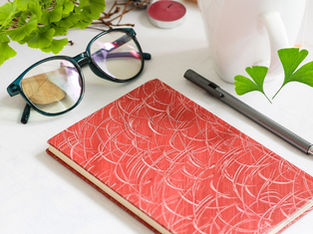Glasses, a red patterned notebook, and a pen rest on a white table. Ginkgo Biloba tree, green Ginkgo Biloba leaves, and a pink candle create a calm setting. Source: Canva