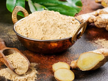 Bowl of ginger powder with a wooden scoop and fresh ginger slices on a rustic surface, green leaves in background, earthy tones. Source: Canva.