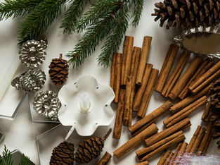 a festive arrangement on a white background, featuring various holiday-themed items. There are several cinnamon sticks bundled together, pinecones, and green pine needles. Additionally, there is a white candle with a decorative holder, a star-shaped cookie cutter, and some metallic star ornaments. The overall composition suggests a cozy, winter holiday scene, possibly related to Christmas.