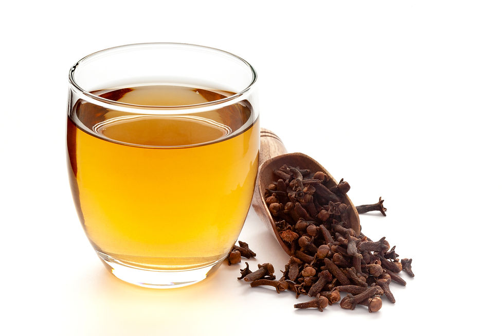 Glass of amber liquid - clove water - next to a wooden scoop with cloves on a white background, creating a warm and aromatic atmosphere. Herbpy Blog.