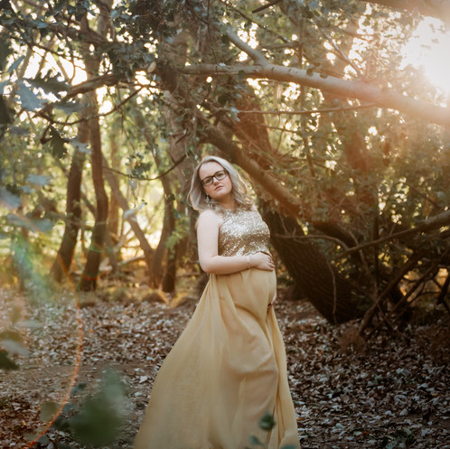 Pregnant woman wearing golden gown with golden sunlight between trees