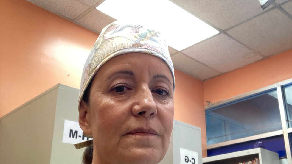 Woman in tan scrubs and floral cap stands in a clinic room by file shelves labeled C-G and A-C, looking serious.