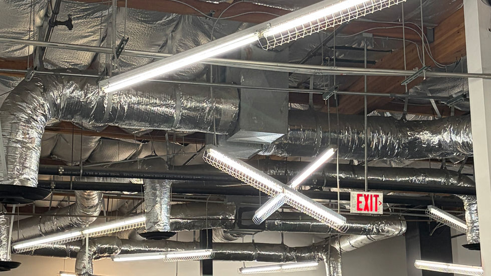 Industrial room with metallic ceiling pipes and bright fluorescent lights. Empty tables and seating, one person sitting. Visible "EXIT" and "two".