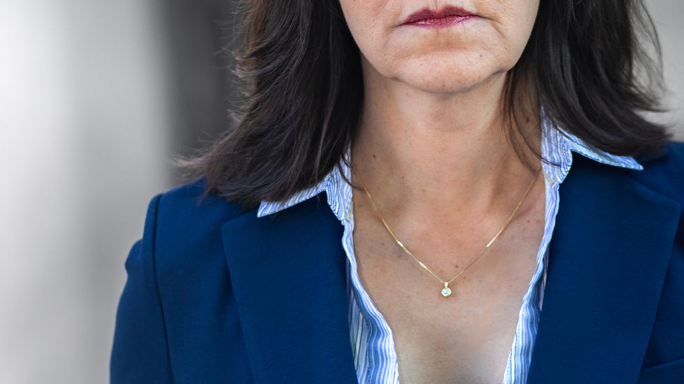Serious woman in a blue blazer and striped shirt stands against a soft gray background, looking directly at the camera.