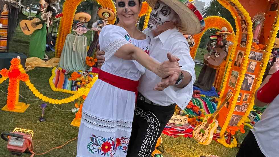 Couple in Day of the Dead makeup dancing, surrounded by colorful altars and marigold decorations, with musicians in the background.