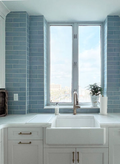 A farmhouse kitchen sink in front of a window with a view to the beach, marble countertops and blue tile